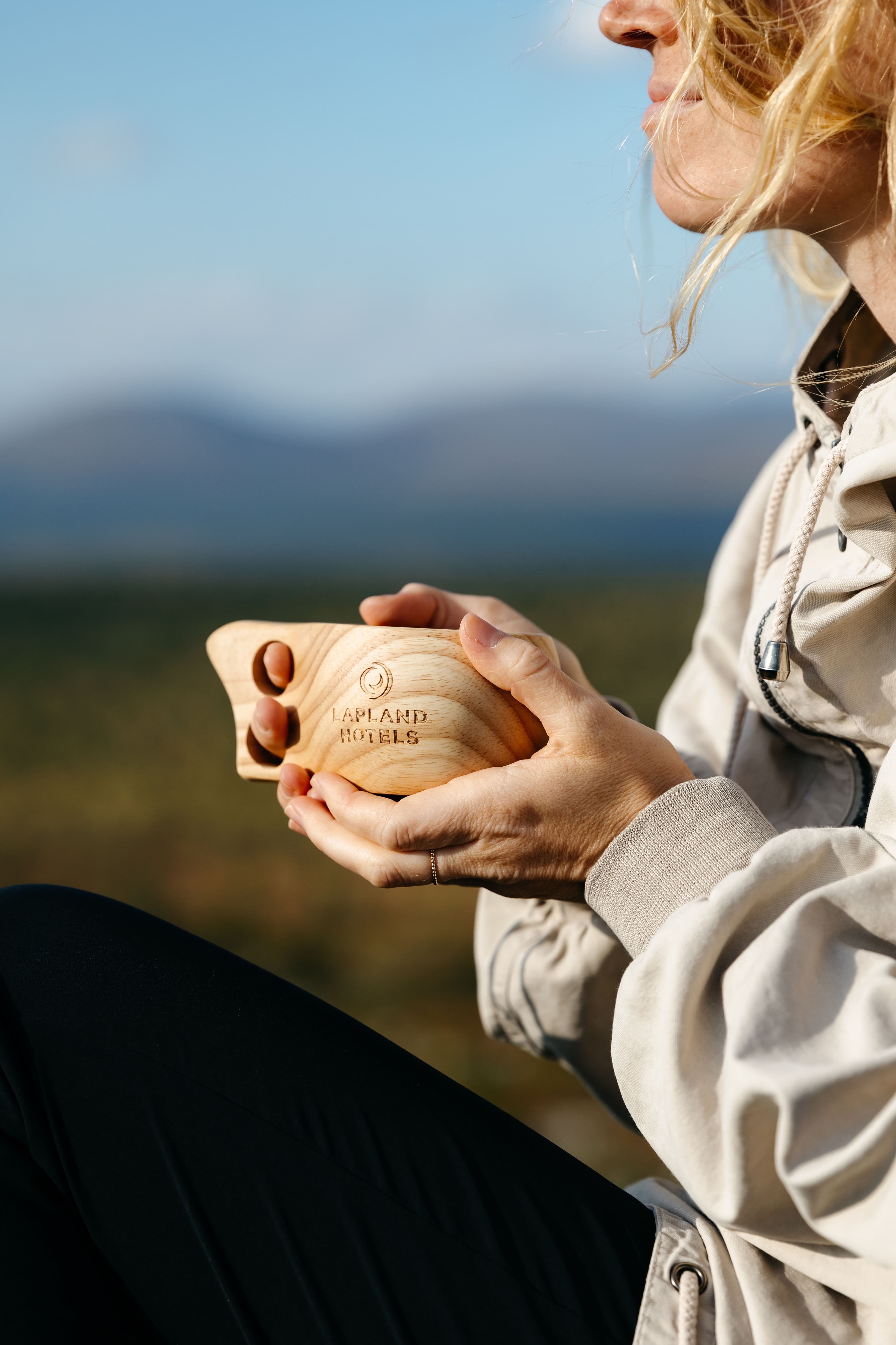 Nainen puinen Lapland Hotels -kuksa kädessä, taustalla avautuvat tunturimaisemat.