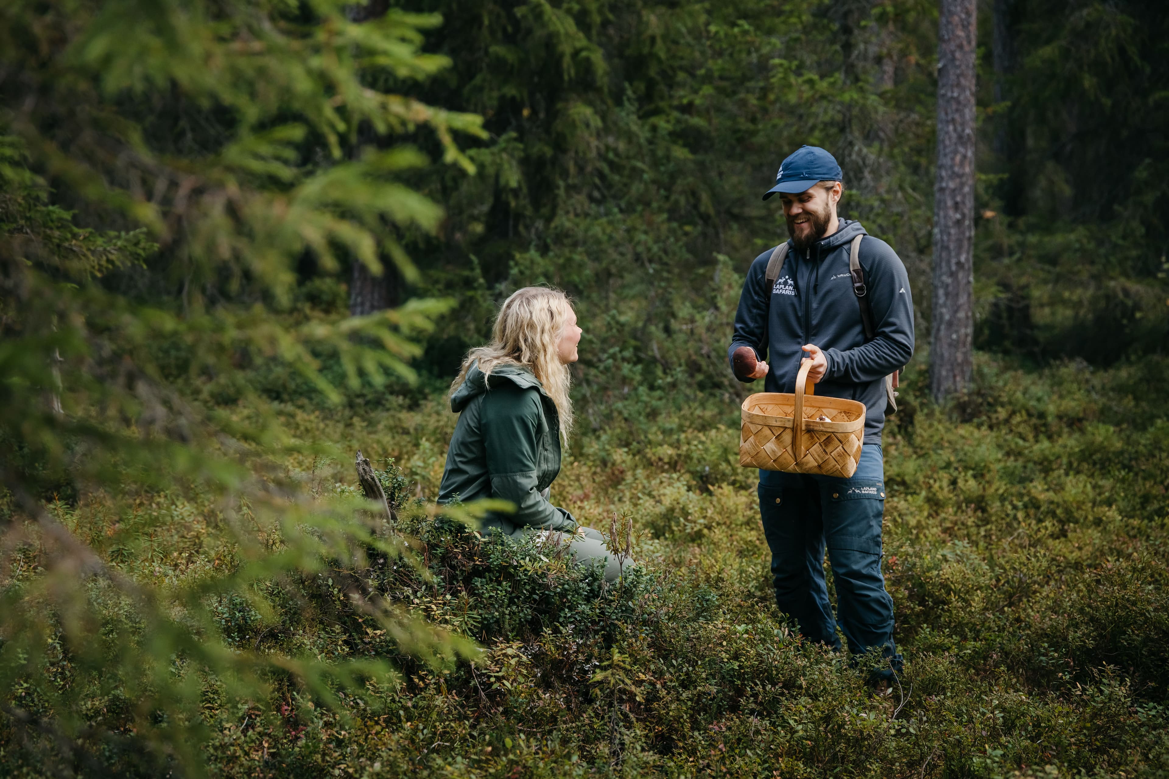 Kaksi henkilöä sienestää metsässä. Toisella on kori kädessään, toinen istuu metsässä puiden keskellä.